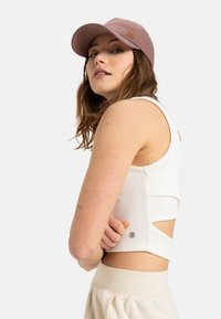 White ribbed crop top with cut-out sides and silver hardware detail, paired with beige high-waisted pants and a lavender cap.