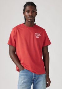 Crvena majica kratkih rukava od pamuka. Sadrži bijeli grafički tekst "HOUSE PLANTS" i "Levi's." Uparena s svijetloplavim poderanim trapericama.