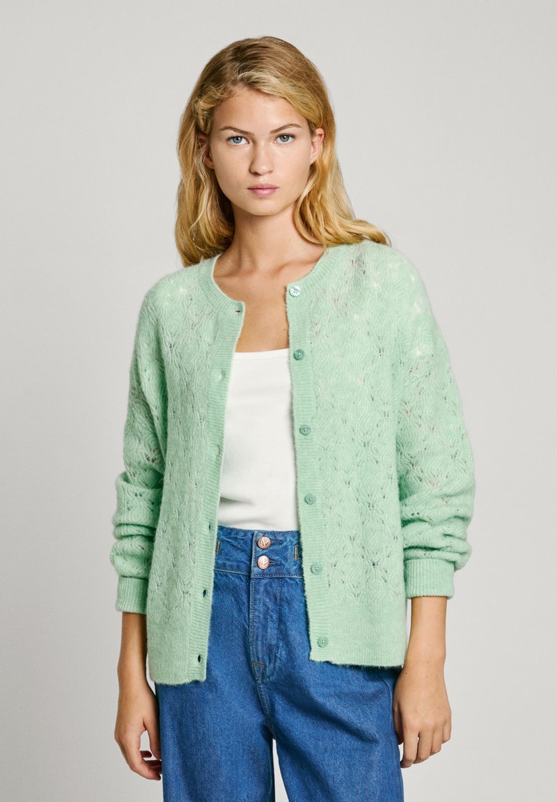 Gaiši zaļš adīts kardigans ar mežģīņu rakstu, ribotām piedurknēm un apaļu dekoltē. Savienots ar baltu topu un ziliem džinsiem.