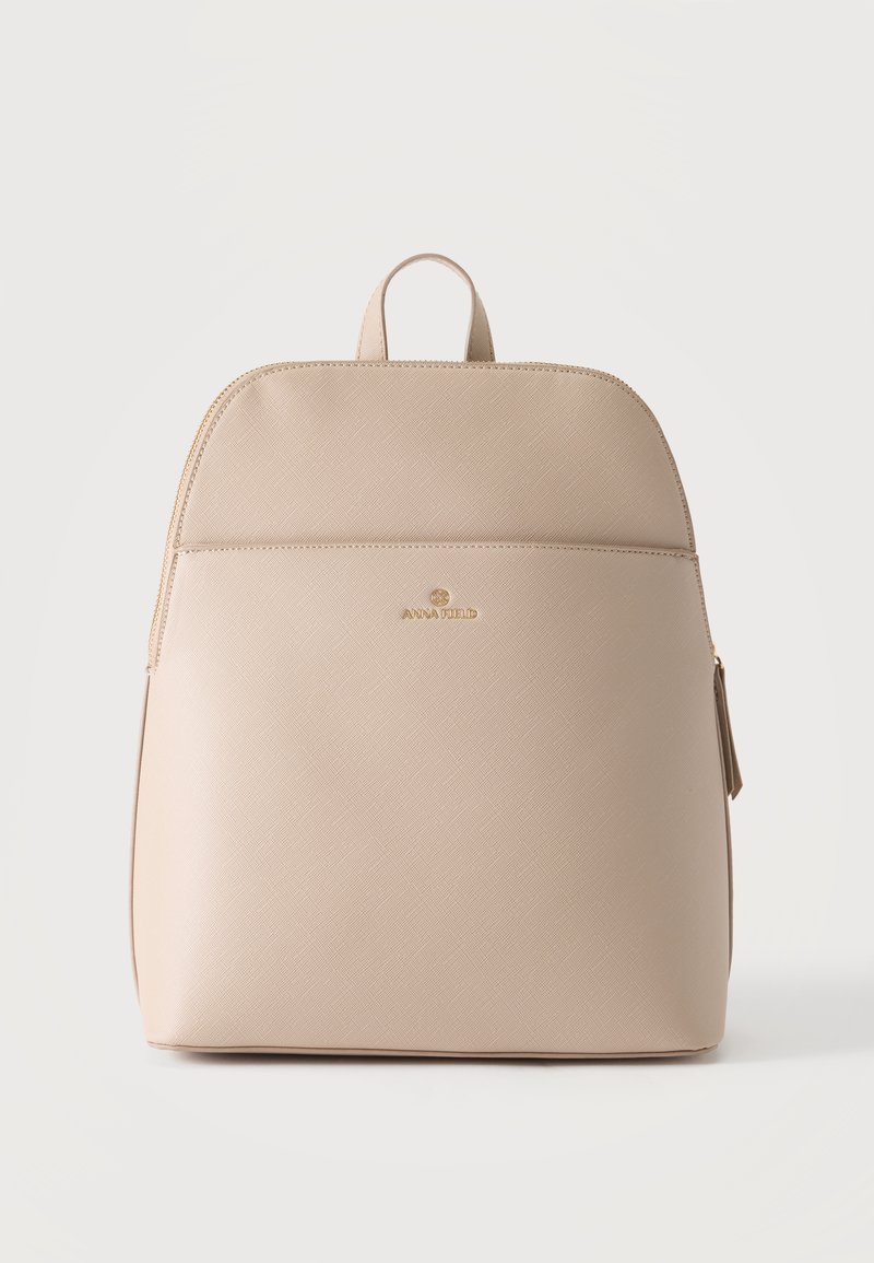 Sac à dos beige texturé avec poignée supérieure, poche avant et logo de marque doré sur un fond clair uni.