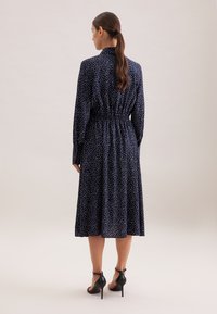 Vestito blu con pois bianchi, maniche lunghe, vita elastica e gonna di lunghezza media. Indossato con sandali neri con tacco alto.