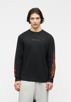Junger Mann trägt ein schwarzes Langarmshirt mit roten Flammengrafiken auf den Ärmeln und einem verblassten Oakley-Logo auf der Brust, kombiniert mit hellgrauen Hosen.