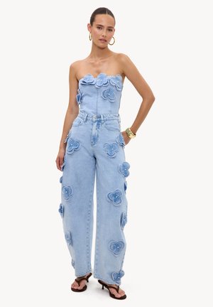 Femme portant une combinaison en denim bleu clair sans bretelles avec des applications florales 3D, des boucles d’oreilles créoles dorées, un bracelet et des sandales noires à talons.
