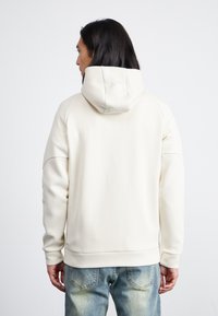Kremfarget hettegenser med avslappet passform, myk tekstur og ribbede ermer, sammen med lyseblå denimjeans.