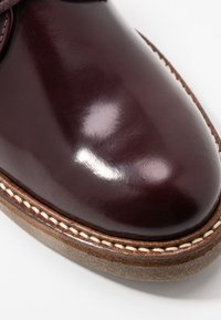 Burgunderfarbene Lederschuhe mit glatter Oberfläche, ausgestattet mit einer braunen Wildledersohle und detaillierter weißer Naht entlang des äußeren Randes.