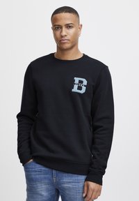 Felpa nera in misto cotone con collo rotondo, polsini a costine e logo blu chiaro con una 'D' sul petto. Indossata con jeans blu.