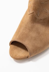 Åben-tå brun ruskindsandal med en syet kant og dekorativ bladvorm på den øverste del. Blød tekstur og ergonomisk design.