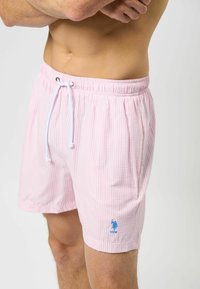Lyse rosa striped badeshorts med elastisk linning, snøring, og en liten blå logo nederst til venstre.