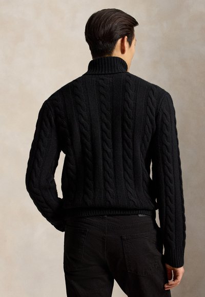Homme portant un pull à col roulé en maille torsadée noire et un pantalon noir, debout dos à la caméra sur un fond neutre.