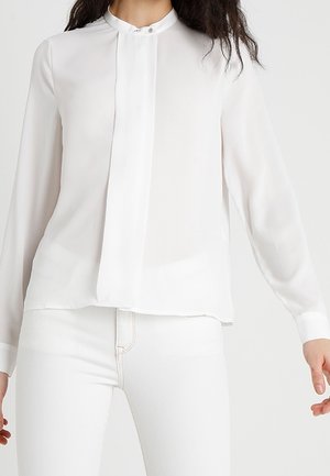Woman wearing a long-sleeve white blouse with a band collar and white fitted pants against a plain background.