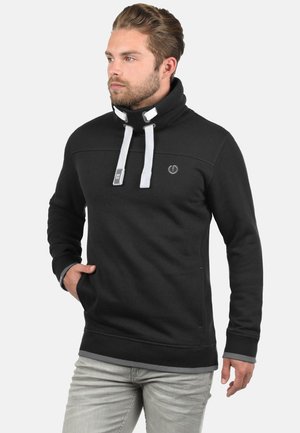 Schwarzer Pullover mit hohem Kragen, weißen Kordeln, Seitentaschen und einem Logo auf der Brust. Obermaterial aus Baumwollmischung mit strukturierter Oberfläche.