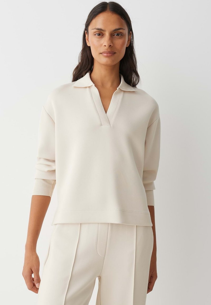 Lichtbeige trui met V-hals en driekwart mouwen, gemaakt van een glad textiel. Heeft geribbelde accenten bij de kraag en manchetten.