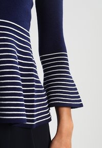 Haut tricoté bleu marine avec des rayures horizontales blanches ; doté de manches évasées et d'une silhouette péplum, mettant en valeur une finition texturée.