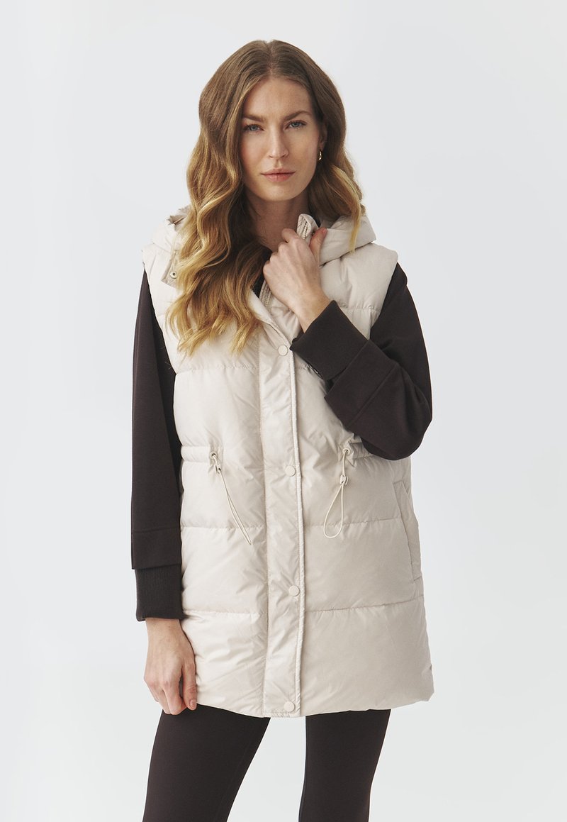 Beige puffer vest with a hood, front snaps, adjustable drawstrings at the waist, and a smooth, shiny texture. Layered over a dark sweatshirt.