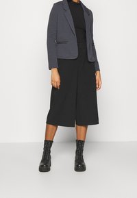 Blazer gris texturé à revers crantés sur un haut noir, associé à un pantalon large noir type culotte et des bottines à lacets noires.