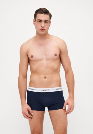 Marineblauwe boxershorts met een witte tailleband waarop het Lacoste-logo is gedrukt. De stof lijkt glad met een aansluitend ontwerp.