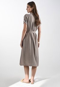 Una donna con capelli lunghi indossa un vestito grigio con cintura, in piedi su uno sfondo chiaro, mostrando uno stile casual ed elegante.