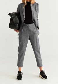 Costume gris en mélange de laine avec veste ajustée, pantalon assorti longueur cheville, et haut noir côtelé. Les accessoires comprennent un grand sac noir et des baskets noires.