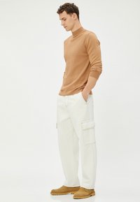 Tan geribbelde trui met hoge hals, gecombineerd met witte cargobroek met zijzakken. Bruine instappers maken de outfit compleet.