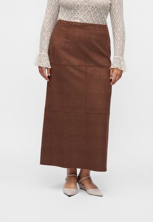 Kvinde iført et langt brunt tekstureret skørt, hvid blonder med lange ærmer og beige flade spidse sko med ankelrem.