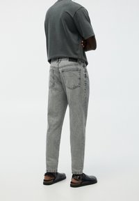 Homme à la peau foncée portant un jean gris délavé, des chaussures noires ouvertes à l'arrière, et une chemise à manches courtes gris foncé, debout les bras croisés.