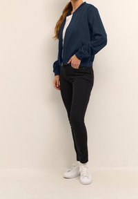 Veste bomber bleu marine, fermeture éclair, poignets et ourlet élastiques, associée à un jean noir et des baskets blanches. Design simple et épuré.
