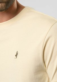 Tricou din bumbac galben deschis cu decolteu clasic și un mic detaliu brodat pe partea stângă a pieptului. Textură moale, design casual.