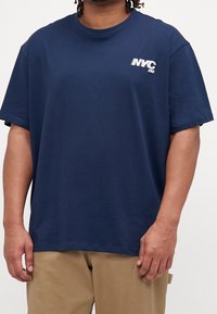 Tmavě modré bavlněné tričko s kulatým výstřihem, krátkými rukávy a bílým nápisem "NYC" na levé straně hrudi. Měkká textura, volný střih.