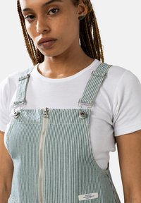 Salopette en denim rayé bleu clair et blanc avec une fermeture éclair sur le devant, des bretelles métalliques réglables, et un patch logo sur le bas à gauche.