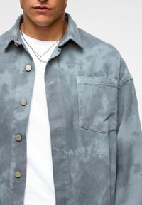 Lichtgrijze, gestructureerde denim shirt met een borstzak, metalen knopen en een ontspannen pasvorm, laag over een wit t-shirt.