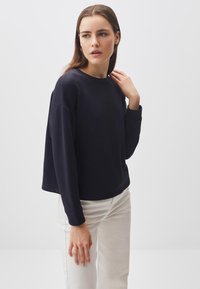 Felpa nera a maniche lunghe con scollo rotondo e vestibilità comoda, abbinata a pantaloni beige. Presenta un tessuto morbido e polsini a coste per una texture aggiuntiva.