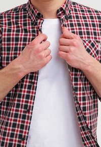 Kädet napittamassa punaista, mustaa ja valkoista ruudullista paitaa yksinkertaisen valkoisen T-paidan päälle henkilön yllä, neutraalilla taustalla.