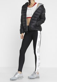 Svart puffjacka med grå huva, kombinerad med svarta leggings med vit FILA-logotyp, och grå träningsskor med strukturerad sula.