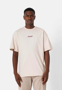 Beżowy, oversize t-shirt wykonany z bawełny, z krótkimi rękawami i okrągłym dekoltem. Z przodu centralnie umieszczone logo w ciemnoczerwonym kolorze.