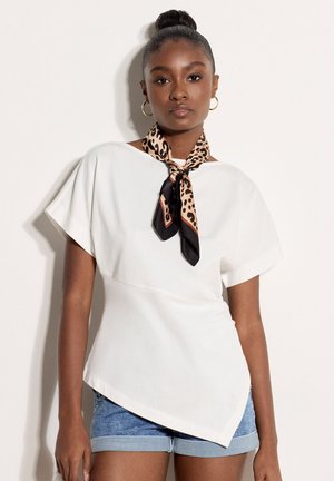 Mujer con piel oscura y cabello recogido en un moño, lleva pendientes de aro dorados, camiseta blanca asimétrica de manga corta, shorts azules remangados y bufanda con estampado de leopardo.
