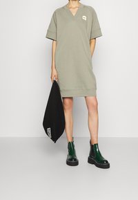 Khaki oversize sukienka-sweterek z dekoltem w serek, krótkimi rękawami i małym logo; zestawiona z zielonymi botkami typu chelsea i czarną torbą na ramię.