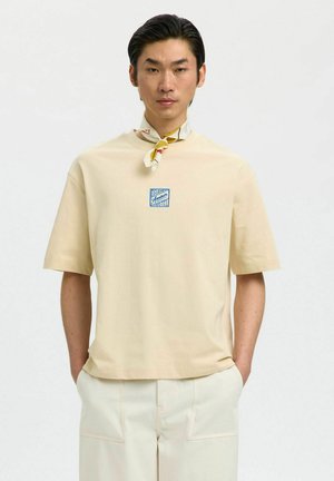 Muž v béžovej košeli s krátkym rukávom, s malým modrým logom, v svetlých nohaviciach, ruky v vreckách a so vzorovaným šálom zviazaným okolo krku.