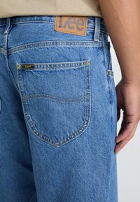 Suurendatud vaade sinistest denimpükside tagataskust, millel on Lee brändi silt, ja isiku parem käsi puusa kõrval.