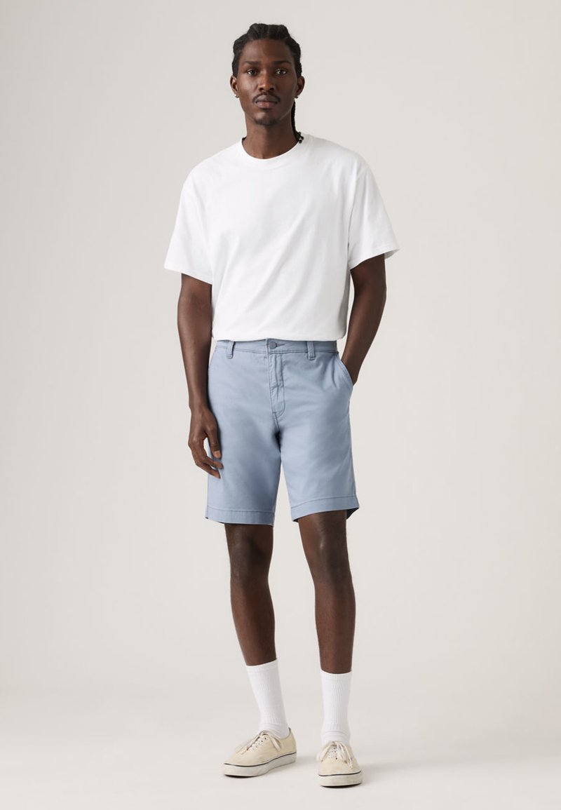 Homme aux cheveux tressés portant un t-shirt blanc, un short bleu clair, des chaussettes blanches et des baskets beiges, debout devant un fond uni.