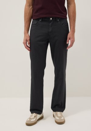 Mann trägt dunkelgraue straight-leg Jeans, burgunderrotes Hemd und beige Sneakers, steht vor neutralem Hintergrund.