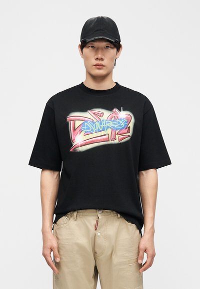 Νεαρό άτομο που φοράει μαύρο καπέλο και μαύρο γραφικό T-shirt με πολύχρωμο σχέδιο σε στυλ γκράφιτι, μπεζ παντελόνι, στέκεται μπροστά σε απλό φόντο.