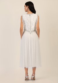 Hvid blondekjole med bølgede ærmer, en tætsiddende bodice og en semi-gennemsigtig polka dot-nederdel; har en baglynlås og teksturerede detaljer.