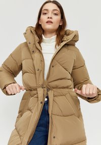 Puffy beige long coat with a hood, zip closure, and drawstring waist. Features diagonal stitching and side pockets. Smooth, insulated fabric.