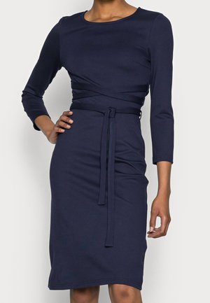 Frau trägt ein tailliertes, knielanges Kleid in Marineblau mit Dreiviertelärmeln und einem gebundenen Taillengürtel.