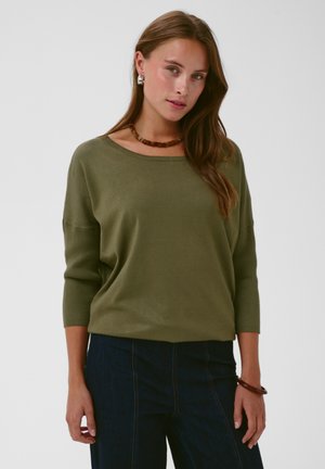 Kvinde med langt brunt hår iført en olivengrøn sweater og mørke bukser, accessoriseret med en brun halskæde, øreringe og armbånd.