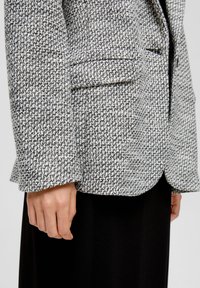 Gewebter Blazer in Schwarz und Weiß, mit einem strukturierten Hahnentrittmuster, strukturiertem Kragen und zwei vorderen Taschen.