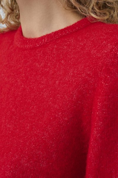 Gros plan sur une personne portant un pull tricoté rouge vif et duveteux avec un col rond, ses cheveux bouclés châtain clair sont visibles.