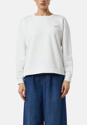 Woman wearing a plain white long-sleeve sweatshirt with small red "Xoxo" text on chest and loose blue denim pants.