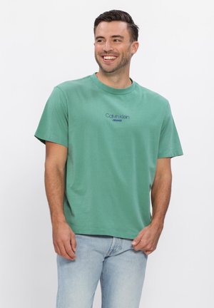 Glimlachende man met donker haar, gekleed in een groen Calvin Klein Jeans T-shirt en lichtblauwe spijkerbroek, staand tegen een effen witte achtergrond.