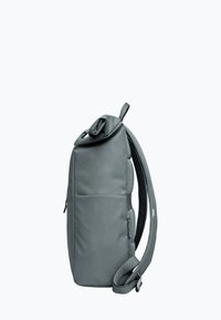 Sac à dos à rabat en gris pâle, avec une surface texturée. Il dispose de bretelles réglables et rembourrées, ainsi que d'une poche zippée latérale pour le rangement.